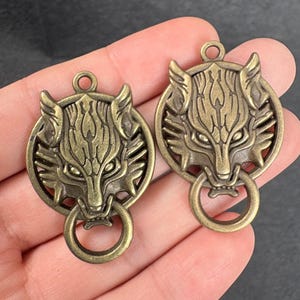 Könnte beinhalten: Zwei bronzefarbene Wolfskopf-Anhänger mit einem Ring am unteren Ende. Das Gesicht des Wolfes ist detailliert mit Fell und Gesichtszügen, eingebettet in einen kreisförmigen Rahmen. Die Anhänger haben oben eine Schlaufe zum Aufhängen.