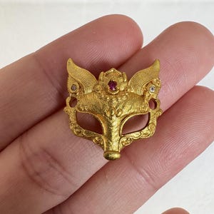 Puede incluir: Colgante de máscara de zorro dorado con detalles intrincados. La máscara presenta una gema de rubí en el centro y dos gemas claras más pequeñas. El colgante tiene una superficie texturizada y está diseñado con elementos en forma de alas.