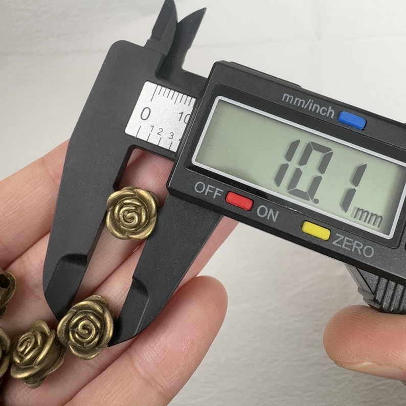 May include: A close-up of a digital caliper measuring a small, antique brass-colored rose. The digital display reads 10.1 mm, indicating the rose's diameter. Several other rose-shaped beads are visible in the background.