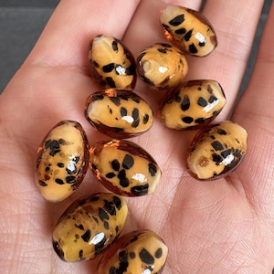 Puede incluir: Un conjunto de cuentas ovaladas con un patrón moteado. Las cuentas son de color ámbar translúcido con manchas marrones oscuras y parecen estar hechas de vidrio o resina. Las cuentas son lisas y brillantes.