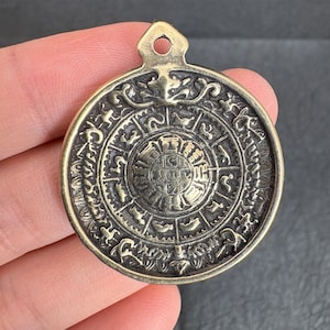 Puede incluir: Un colgante circular de color bronce con intrincados diseños en relieve. El colgante presenta un emblema central rodeado de patrones y símbolos detallados. Tiene un pequeño lazo en la parte superior para colgar, probablemente una joya o un objeto decorativo.