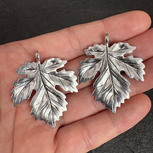 Può includere: Due ciondoli in metallo color argento a forma di foglie di vite. Ogni ciondolo presenta un motivo a venature dettagliato e in rilievo sulla superficie e un piccolo occhiello circolare sulla parte superiore per l'aggancio. Il metallo ha una finitura lucida con aree incavate scure.