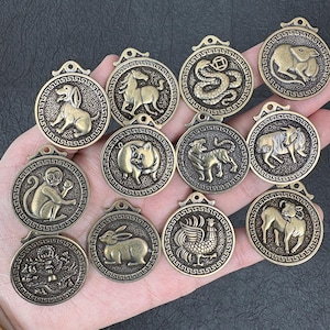 Może przedstawiać: Kolekcja brązowych wisiorków zodiakalnych, każdy z innym symbolem zwierzęcym w okrągłej ramie. Wisiorki są trzymane w dłoni, prezentując szczegółowe rzeźby reliefowe każdego zwierzęcia.