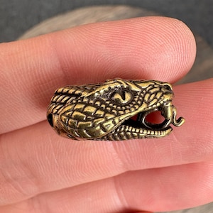 May include: A small, metallic, brass-colored bead shaped like a snake head. The bead features detailed, raised scale textures across the surface, a prominent eye, and an open mouth with a visible, protruding tongue. The object is compact and cylindrical.