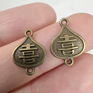 May include: Two antique bronze-colored charms. Each charm has a teardrop shape with a loop at the top and a smaller loop at the bottom. The charms feature a Chinese character design.