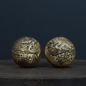 Pode incluir: Duas esferas antigas de latão com intrincados desenhos de dragões e nuvens. As esferas apresentam padrões em espiral e um acabamento texturizado, repousando sobre uma superfície de madeira escura. As esferas são de cor dourada quente.