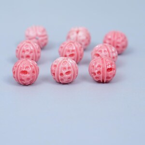May include: A collection of pink, spherical beads with a carved, textured surface. The beads are arranged on a light blue background. The beads have a series of small, evenly spaced holes.