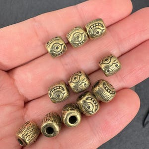 May include: A collection of antique brass-colored beads with intricate engraved designs. The beads are square and cylindrical, featuring eye-like and geometric patterns. The beads are displayed in a hand, showcasing their detailed craftsmanship.
