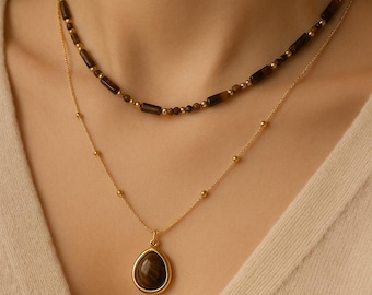 May include: A layered necklace set featuring a gold chain with a teardrop pendant and a beaded necklace. The pendant has a brown stone. The second necklace has brown rectangular beads and small gold beads.