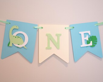 Könnte beinhalten: Ein Banner mit dem Wort "ONE" in grünen und weißen Buchstaben. Das Banner besteht aus drei Wimpeln in Hellblau und Creme. Der erste Wimpel hat einen grünen Dinosaurier und den Buchstaben "O". Der dritte Wimpel hat ein grünes Blatt und den Buchstaben "E".