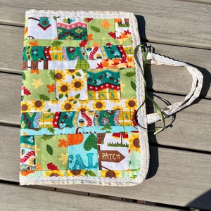 May include: A quilted journal cover with a patchwork design featuring sunflowers, pumpkins, and fall leaves. The cover has a white lace trim and a fabric handle with green ribbon accents. The words "FALL PATCH" are visible on the cover.