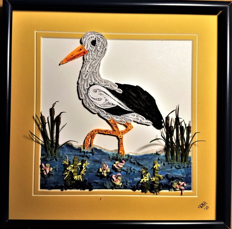 May include: A white and black paper quilled stork standing in a blue water scene with green reeds. The artwork is framed in a yellow and black frame.