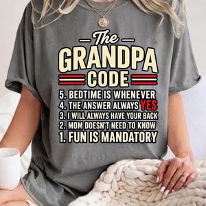 May include: A charcoal gray t-shirt featuring the text The GRANDPA CODE followed by a numbered list: 5. BEDTIME IS WHENEVER, 4. THE ANSWER ALWAYS YES, 3. I WILL ALWAYS HAVE YOUR BACK, 2. MOM DOESN'T NEED TO KNOW, 1. FUN IS MANDATORY. The text is in black and cream.
