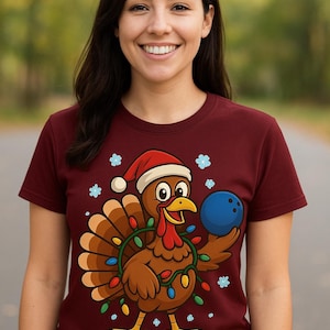 Puede incluir: Camiseta burdeos con un pavo de dibujos animados que lleva un gorro de Papá Noel y sostiene una bola de bolos azul. El pavo está decorado con luces navideñas. La camiseta tiene cuello redondo y mangas cortas.