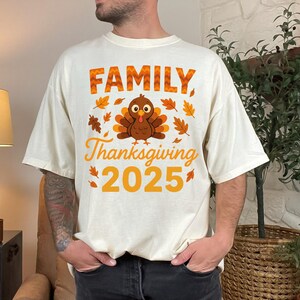 Pode incluir: Camiseta creme com tema de Ação de Graças. A camiseta apresenta as palavras "FAMILY Thanksgiving 2025" em laranja, junto com um desenho de peru e folhas de outono. A camiseta tem um corte folgado.