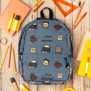 Peut inclure: Un sac à dos bleu avec un motif de visages tristes, de tasses à café et d'imprimantes, avec le texte "... Over Worked". L'image comprend des fournitures scolaires comme des crayons, des surligneurs et des cahiers, sur une surface en bois.