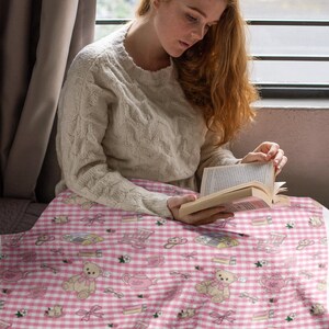 May include: A pink and white checkered blanket with a repeating pattern of teddy bears, teapots, and bows. The blanket is draped over a person reading a book. The person is wearing a cream-colored sweater.