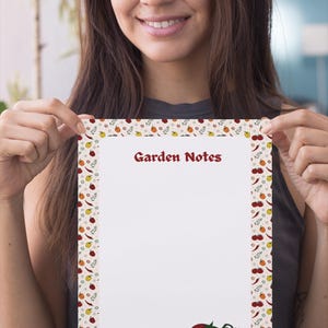 Könnte beinhalten: Ein rechteckiger Notizblock mit weißer Mitte und einem Rand aus kleinen Illustrationen von roten Tomaten, gelben Paprika und grünen Kräutern. Der Text Garden Notes ist oben in Rot gedruckt, zwei rote Tomaten befinden sich unten rechts.