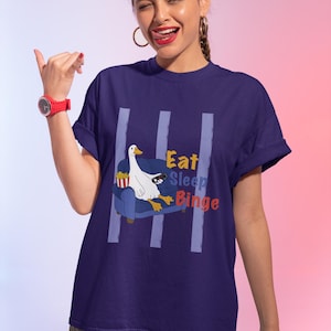 Könnte beinhalten: Marineblaues T-Shirt mit einer Grafik einer weißen Gans auf einem blauen Sofa, die eine Fernbedienung und Popcorn hält. Der Text "Eat Sleep Binge" ist in Gelb und Rot. Vertikale hellblaue Streifen sind auf dem Shirt.