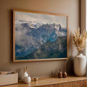 May include: A framed landscape print of a mountain range with snow-capped peaks, shrouded in mist. The artwork is displayed on a neutral-colored wall above a wooden cabinet. Decorative vases and a woven basket add to the room's decor.