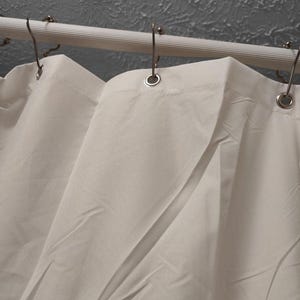 May include: A white shower curtain with metal eyelets hangs from a white, ribbed shower rod. The curtain is made of a smooth, opaque fabric and is partially draped, showing folds and shadows. The background is a textured grey wall.