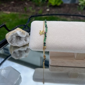 May include: A gold-colored bracelet with green and black beads and a small butterfly charm. The bracelet is displayed on a white, rectangular stand. The background includes a blurred view of greenery and a reflective surface.