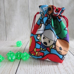 May include: A red drawstring bag with a colorful cartoon print of superheroes. The bag is filled with green dice.