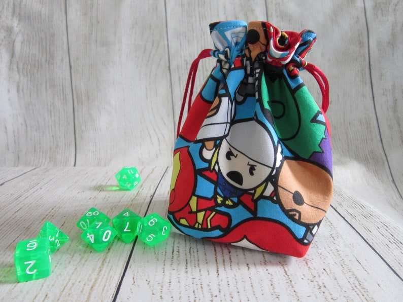 May include: A red drawstring bag with a colorful cartoon print of superheroes. The bag is filled with green dice.
