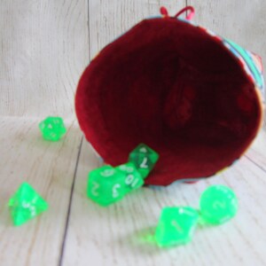 May include: A red fabric dice bag with a colorful pattern is tipped over, spilling out green dice with white numbers.