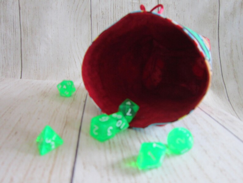 May include: A red fabric dice bag with a colorful pattern is tipped over, spilling out green dice with white numbers.