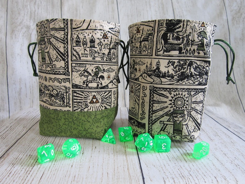May include: Two green fabric dice bags with a white and black print of a video game character. The bags have a drawstring closure. Green dice are scattered in front of the bags.
