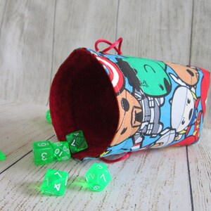 May include: A red and blue dice bag with a cartoon print of superheroes. The bag is open and green dice are spilling out.