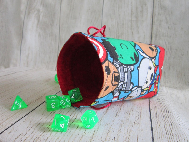 May include: A red and blue dice bag with a cartoon print of superheroes. The bag is open and green dice are spilling out.