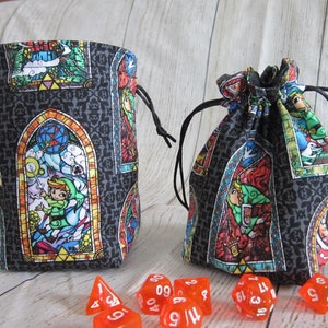 May include: Two black fabric dice bags with a stained glass pattern featuring a video game character. The bags have drawstrings and are filled with red dice.