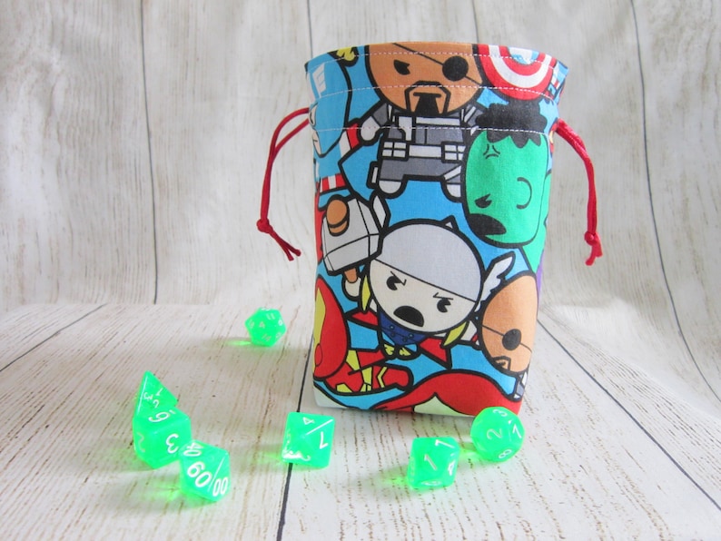 May include: A red drawstring bag with a blue background and a pattern of cartoon superheroes. The bag is filled with green dice.