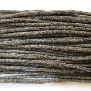 Könnte beinhalten: Nahaufnahme von mehreren Strähnen grauer Dreadlock-Extensions. Die Dreadlocks sind dick, zylindrisch und haben ein strukturiertes, faseriges Aussehen. Die Farbe ist ein gedämpftes, erdiges Grau, der Hintergrund ist weiß.