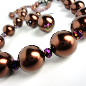 Pode incluir: Um colar de pérolas marrons com detalhes iridescentes. O colar é feito de pérolas grandes e redondas e apresenta pequenos cristais coloridos.