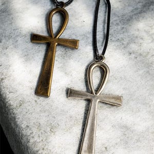 May include: Two Ankh necklaces with black cords. One Ankh is bronze-colored, the other is silver-colored. The Ankh is an ancient Egyptian hieroglyphic symbol representing life. The necklaces are displayed on a textured, light gray surface.