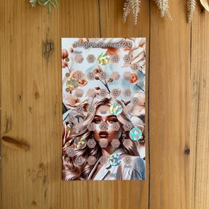 Op de afbeelding: Een rechthoekige kaart met de tekst "Notgroschenweg 2.0" en een bloemenachtergrond. De kaart toont een portret van een vrouw en verschillende holografische stickers met nummers. De kaart ligt op een houten oppervlak.