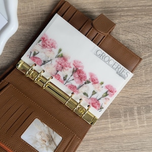Puede incluir: Un planificador de cuero marrón con anillos dorados, que contiene una hoja de presupuesto con flores rosas y la etiqueta "GROCERIES". Una pequeña tarjeta con texto es visible. El planificador está abierto, revelando ranuras para tarjetas.
