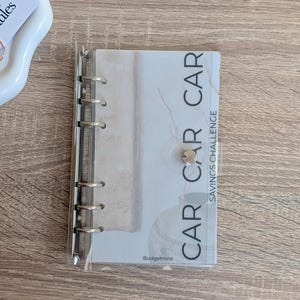 Può includere: Un'agenda trasparente ad anelli con le parole "CAR CAR" e "SAVINGS CHALLENGE" stampate sulla copertina. L'agenda è aperta, rivelando pagine a righe. Lo sfondo è un legno chiaro.