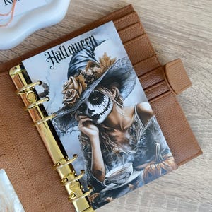 Könnte beinhalten: Ein brauner Lederplaner mit goldfarbenen Ringen enthält einen dekorativen Einsatz. Der Einsatz zeigt eine Frau mit Halloween-Make-up und einem Hexenhut, mit dem Wort "Halloween" oben. Eine Teetasse und ein Kürbis sind ebenfalls zu sehen.