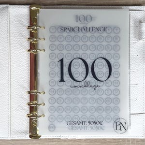 May include: A white binder with gold hardware holds a clear sheet with a 100-circle savings challenge. The text "100 Sparchallenge" and "100 umschläge" are visible. The total savings amount is 5050€.