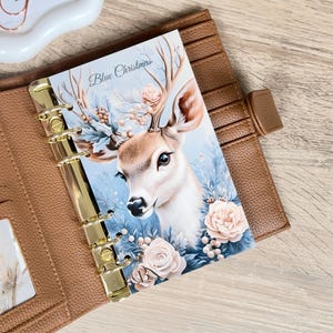 Op de afbeelding: Een bruine leren planner met gouden hardware, opengeklapt op een pagina met een hertenillustratie. De illustratie toont een hert met geweien, omringd door blauwe en roze bloemelementen, en de tekst "Blue Christmas".