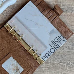 Könnte beinhalten: Ein brauner Lederplaner mit goldfarbenen Ringen und einer transparenten Kunststoffauflage. Die Auflage hat ein weißes und goldenes Design und die Worte "HIGH PRIORITY" sind aufgedruckt. Der Planer ist geöffnet und zeigt Taschen und einen dekorativen Einsatz.
