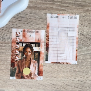 Puede incluir: Dos tarjetas sobre una superficie de madera. Una tarjeta presenta a una mujer con una blazer rosa, con el texto "MINI GROSCHEN" y números. La otra tarjeta tiene el texto "datum +/- summe" y una cuadrícula.