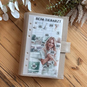 Könnte beinhalten: Ein beiger Planer mit strukturierter Hülle und Druckknopfverschluss. Der Planer hat eine transparente Folie mit dem Text "BEIM TIERARZT" und einem Raster mit Zahlen und Tierbildern. Eine Frau hält eine Katze in der Mitte.