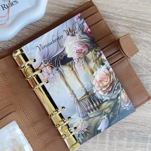 Può includere: Un'agenda in pelle marrone con anelli dorati e un inserto decorativo. L'inserto presenta un motivo floreale e il testo "Verzauberter Wald". L'agenda è aperta, rivelando fessure per carte e una tasca trasparente.