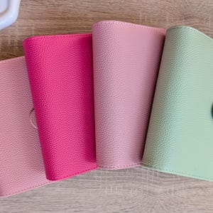 Puede incluir: Cuatro cuadernos de colores pastel con textura de cuero. Los colores son rosa claro, rosa intenso, rosa medio y verde claro. El cuaderno verde claro tiene un cierre de lengüeta. Los cuadernos están colocados sobre una superficie de vetas de madera clara.