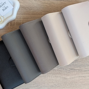 Puede incluir: Cinco planificadores con textura de cuero en diferentes tonos de gris y blanco roto. Los planificadores están cerrados y tienen un cierre de botón. Un plato blanco pequeño con clips dorados y una tarjeta que dice "Your Money Your Rules" está en la esquina superior izquierda.